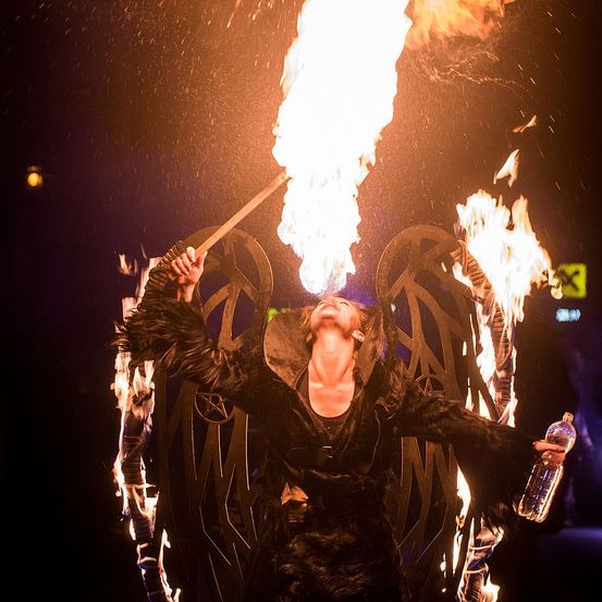 Ein Performer bläst Feuer aus einer Fackel und hält eine Flasche in der anderen Hand. Sie stehen mit dem Rücken zu einem flammenverzierten Hintergrund.