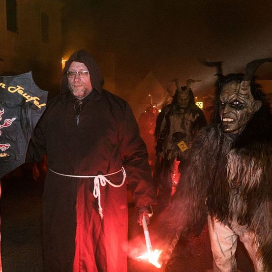 Ein Mann in einem schwarzen Gewand hält eine Fackel mit einer Flamme und steht neben einer Person in einem hörnertragenden Dämonenkostüm.