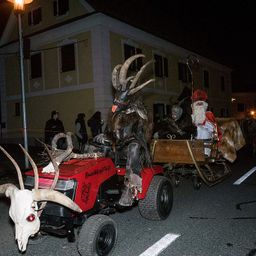 In der Nacht fährt eine Person in einem Ziegenkostüm einen roten Traktor mit einem Schädel an der Vorderseite, während eine andere Person als Weihnachtsmann in einem hölzernen Schlitten sitzt. Ein Gebäude mit Fenstern ist im Hintergrund zu sehen.