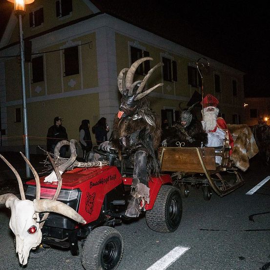 In der Nacht fährt eine Person in einem Ziegenkostüm einen roten Traktor mit einem Schädel an der Vorderseite, während eine andere Person als Weihnachtsmann in einem hölzernen Schlitten sitzt. Ein Gebäude mit Fenstern ist im Hintergrund zu sehen.