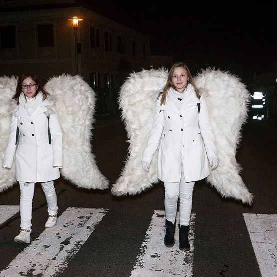 Zwei Frauen, die als Engel gekleidet sind, stehen nachts auf einem Zebrastreifen, eine hält einen Rucksack, die andere trägt eine Brille. Hinter ihnen steht ein Gebäude mit einer Straßenlaterne.