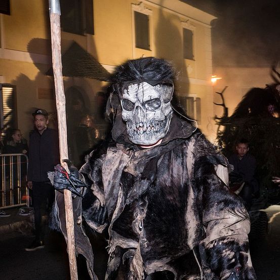 Eine Person mit Totenkopfmaske und einem Stab in der Hand läuft bei Nacht in einer Parade. Zuschauer stehen hinter einem Zaun, Gebäude sind im Hintergrund.
