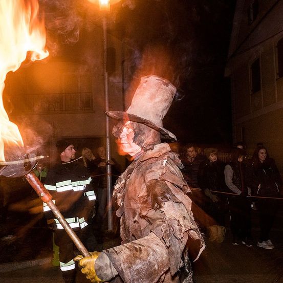 Ein Mann in einem zerlumpten Kostüm mit einem breitkrempigen Hut hält in der Nacht eine flammende Fackel. Ein Feuerwehrmann steht in der Nähe und beobachtet. Zuschauer versammeln sich hinter einem Seilzaun und beobachten die Szene.