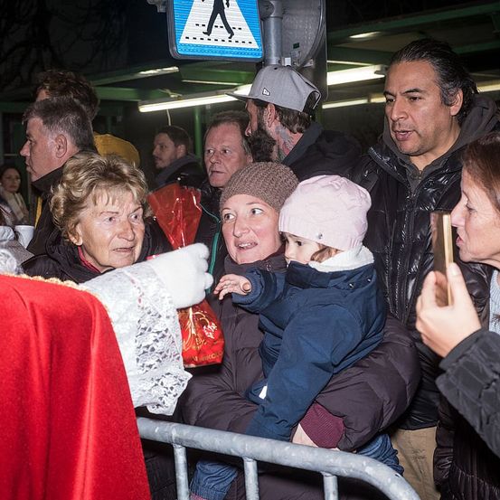 Eine Gruppe von Menschen, einschließlich eines Babys, versammelt sich draußen, möglicherweise in einer Warteschlange. Eine Person in einem roten Tuch hält etwas einer älteren Frau entgegen. Hinter ihnen tragen die Menschen verschiedene Winterkleidung.