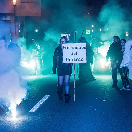 Eine Frau hält ein Schild mit der Aufschrift Hermanos del Infierno in der Hand, während sie die Straße entlanggeht. Die Straße ist mit grünem Rauch bedeckt.