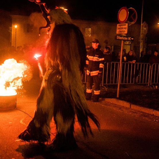 Eine Person in einem Kostüm steht neben einem Feuer und hält eine Fackel. Ein Feuerwehrmann steht in der Nähe, und eine Menschenmenge beobachtet. Es gibt ein Schild mit 'Offentlich' darauf.
