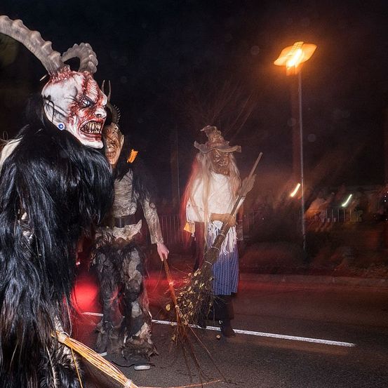Drei Personen in Kostümen stehen nachts auf einer Straße. Einer trägt eine gehörnte Maske, ein anderer einen Hexenhut, und alle halten Besen. Eine Straßenlaterne beleuchtet die Szene.