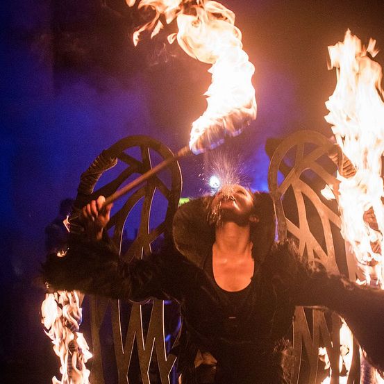 Eine Person führt eine Feuerspuck-Darbietung mit zwei Fackeln vor einer großen Metallstruktur durch.