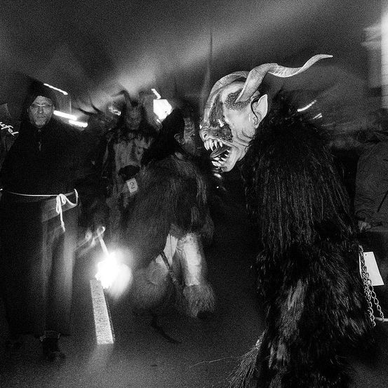 Eine Gruppe von Menschen in Kostümen, einschließlich einer gehörnten Maske, versammelt sich in einem dunklen Bereich. Einige halten brennende Fackeln.