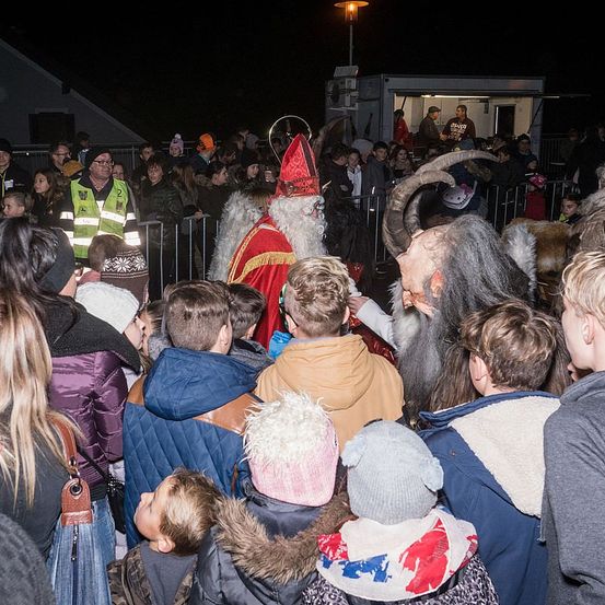 Eine Menschenmenge versammelt sich nachts um eine als Weihnachtsmann verkleidete Person und eine als Krampus, die beide Kinder unterhalten.