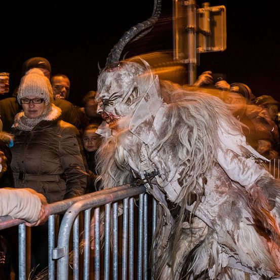 Eine Gruppe von Menschen beobachtet eine Person in einem Dämonenkostüm mit Hörnern und künstlichem Blut, die hinter einer Barriere steht.
