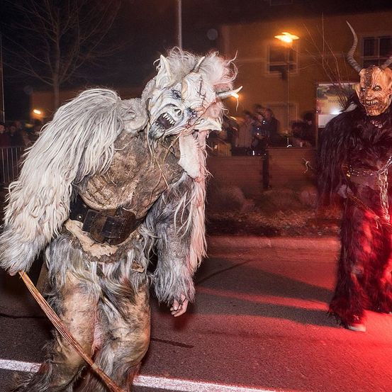 Eine Person in einem hörnertragenden Monsterkostüm geht bei Nacht eine Straße entlang, mit einer weiteren Person in ähnlicher Kleidung hinter ihr. Eine Menge schaut von einer Mauer aus zu.