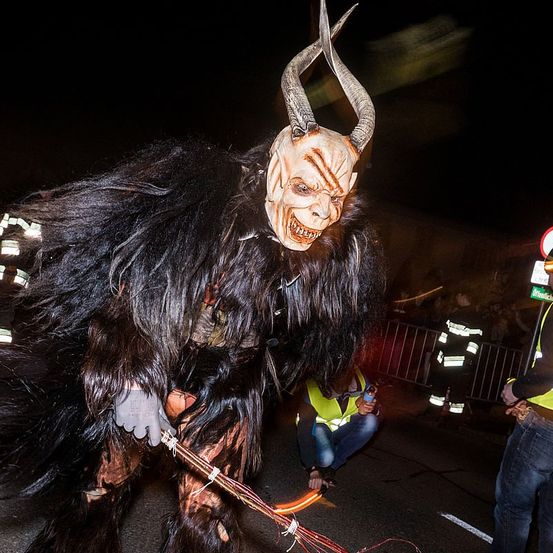 Eine Person in einem gehörnten Kostüm mit einem gruseligen Gesicht steht bei Nacht. Ein Mann in einer gelben Weste kniet in der Nähe. Dahinter ist ein Metallzaun und eine weitere Person in einer reflektierenden Weste.