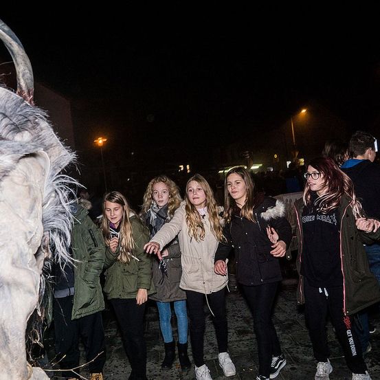 Fünf Mädchen stehen bei Nacht draußen und unterhalten sich lachend. Eine zeigt auf etwas vor ihnen. Sie sind für kaltes Wetter gekleidet, mit Jacken und Stiefeln. Ein großes, pelziges Wesen mit Hörnern steht links von ihnen.