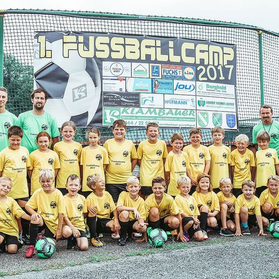 Eine Fußballmannschaft posiert für ein Gruppenfoto vor einem Banner, das '1. Fussballcamp 2017' sagt, mit einem Netzzaun und grünen Bäumen im Hintergrund.