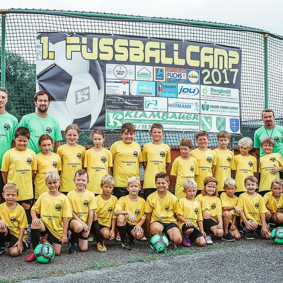 Eine Gruppe von Kindern in gelben Trikots mit verschiedenen Sponsoren-Logos posiert für ein Foto vor einem Fußballnetz. Zwei Männer stehen an den Seiten. Ein Banner hinter ihnen trägt den Text '1. Fussballcamp 2017'.