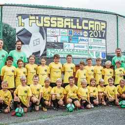Eine Gruppe von Kindern in gelben Trikots mit verschiedenen Sponsoren-Logos posiert für ein Foto vor einem Fußballnetz. Zwei Männer stehen an den Seiten. Ein Banner hinter ihnen trägt den Text '1. Fussballcamp 2017'.