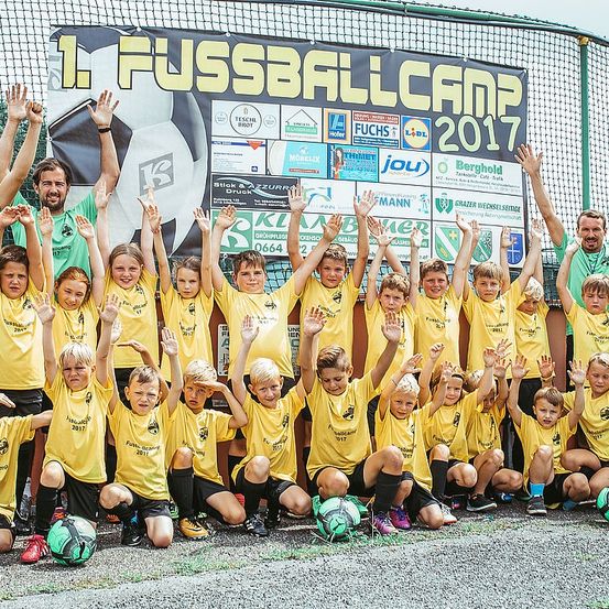 Gruppenfoto von Kindern und Erwachsenen bei einem Fußballcamp, die gelbe Trikots tragen, mit einem Banner im Hintergrund, das 1. Fussballcamp 2017 besagt.