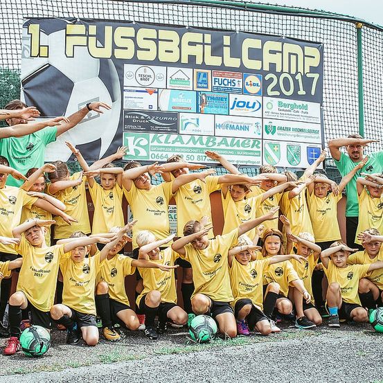Eine Fußballmannschaft posiert für ein Foto, mit den Händen an die Stirn gehalten, um eine Sonnenblende zu simulieren. Sie stehen vor einem Banner, das 1. Fussballcamp 2017 beschriftet ist.