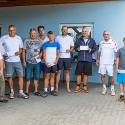 Eine Gruppe von Männern und Jungen steht zusammen, hält Urkunden in den Händen, trägt Freizeitkleidung, mit einer blauen Wand und einem Fenster im Hintergrund.