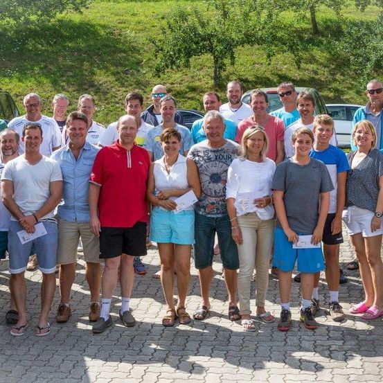 Eine Gruppe von Menschen in lässiger Sommerkleidung steht in einer Reihe und lächelt für ein Foto mit Papieren in den Händen. Dahinter ist ein grün bewachsener Hügel mit geparkten Autos.