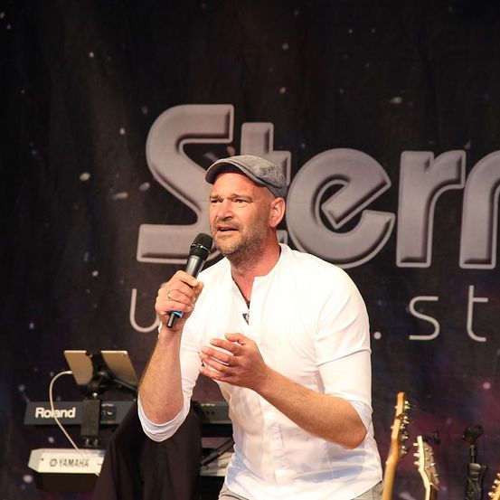 Ein Mann auf der Bühne mit einem Mikrofon, der eine Kappe und ein weißes Hemd trägt, scheint bei einer Musikveranstaltung aufzutreten. Hinter ihm befindet sich ein Banner mit dem Wort 'Stern' und Musikinstrumente auf der Bühne.