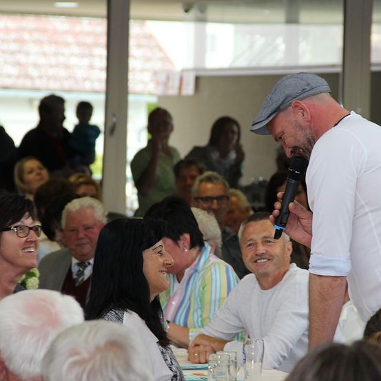 Ein Mann mit einer Kappe spricht in ein Mikrofon, umgeben von einem Publikum aus Erwachsenen, einige lächeln und andere beobachten aufmerksam, mit einer verschwommenen Ansicht von Menschen draußen.