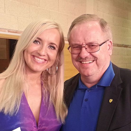 Ein lächelnder Mann und eine Frau posieren für ein Bild. Die Frau trägt ein lila Kleid und große Ohrringe. Der Mann trägt ein blaues Hemd mit einem Logo auf der linken Brust und eine Brille.