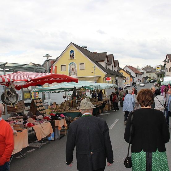 Ein Straßenmarkt mit vielen Ständen, die verschiedene Artikel wie Hüte verkaufen, ist voller Fußgänger. Traditionelle gelbe Häuser und geparkte Autos säumen die Straße.