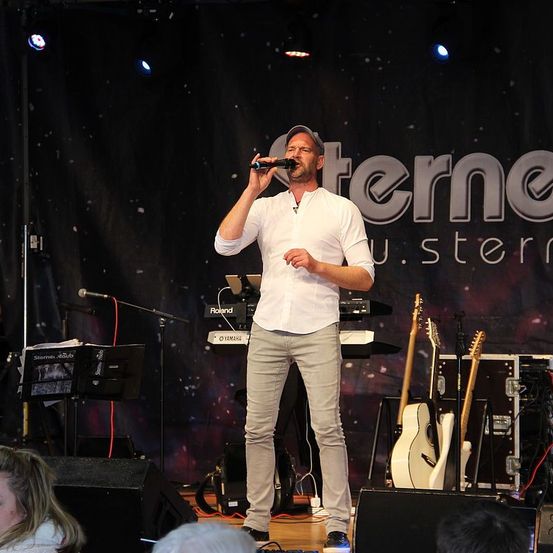 Ein Mann singt auf einer Bühne mit einem Mikrofon, er trägt ein weißes Hemd und beige Hosen. Hinter ihm stehen ein Keyboard und Gitarren, und ein Banner mit dem Wort 'Stern' ist sichtbar. Scheinwerfer sind auf der Bühne montiert.