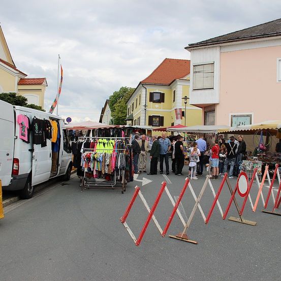 Ein Straßenmarkt mit Verkaufsständen für Kleidung, umgeben von Menschen und Absperrungen an einem bewölkten Tag.