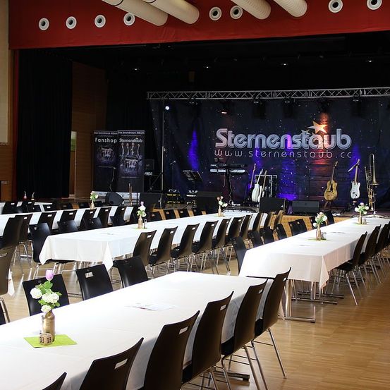 Ein Indoor-Veranstaltungsraum mit festlich gedeckten Tischen. Eine Bühne mit Musikinstrumenten und einem Banner mit der Aufschrift Sternenstaub ist aufgebaut.