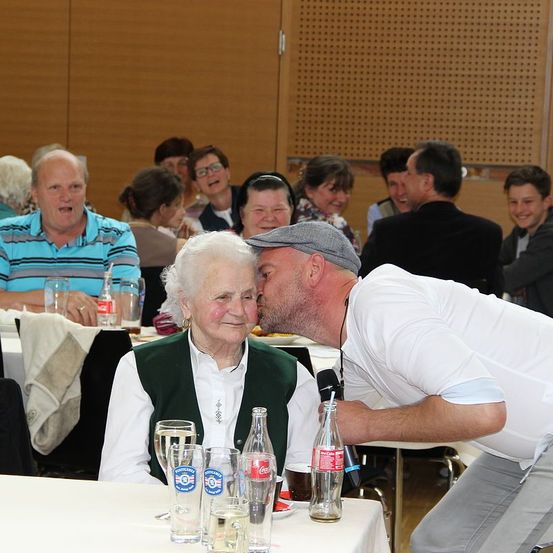 Eine ältere Frau sitzt mit einem Mann, der sich über sie beugt, um sie auf die Wange zu küssen, an einem Tisch. Im Hintergrund lachen und unterhalten sich die Menschen. Gläser und Flaschen stehen auf dem Tisch.