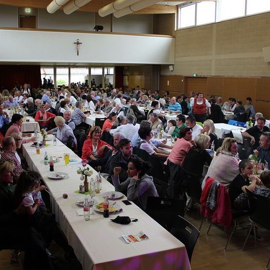 Eine große Gruppe von Menschen sitzt an langen Tischen in einem Bankettsaal. Sie essen. Der Saal hat Holzböden und große Fenster.