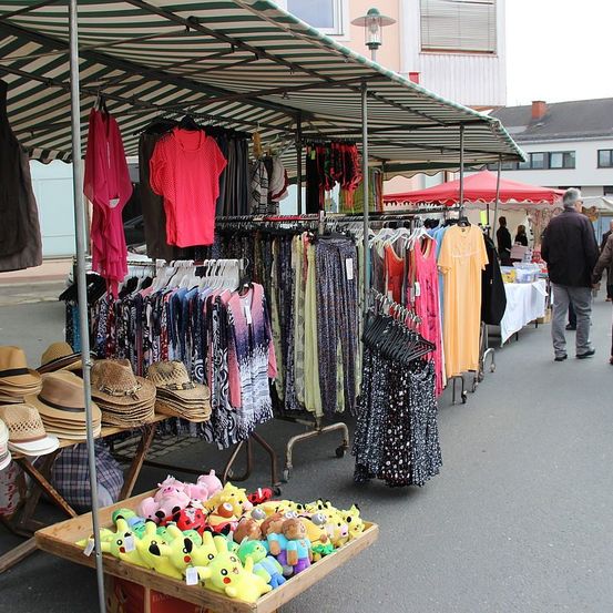 Ein Outdoor-Markt mit verschiedenen Ständen, die Kleidung, Hüte und Spielzeug verkaufen. Mehrere Leute gehen herum, und eine Person steht neben einem Stand.