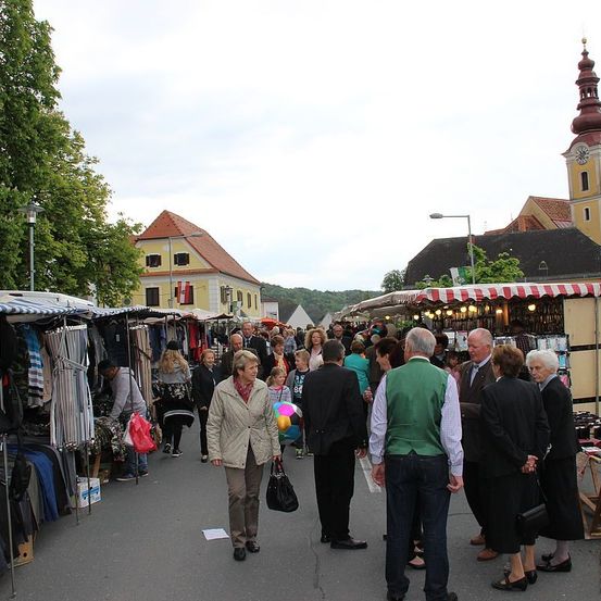 Ein geschäftiger Outdoor-Markt mit verschiedenen Ständen und Menschen, die gehen und plaudern. Der Markt ist unter einem bewölkten Himmel. Im Hintergrund befinden sich Gebäude und ein Turm mit einer Uhr.