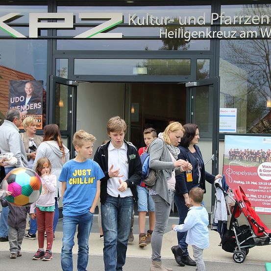 Eine Gruppe von Menschen, einschließlich Kindern und Erwachsenen, versammelt sich vor einem Gebäude mit Glaswänden und Türen. Einige unterhalten sich, während andere Taschen tragen. Ein Kind hält einen bunten Ball.