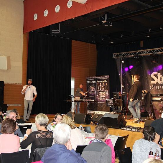 Eine Gruppe von Menschen sitzt in Stühlen vor einer Bühne und schaut sich eine Aufführung an. Ein Mann auf der Bühne singt und hält ein Mikrofon. Ein anderer Mann spielt Gitarre. Dahinter steht ein Tisch mit Laptop, Lautsprecher und einer Flasche.