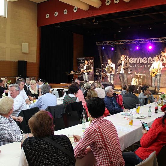 Eine Gruppe von Menschen sitzt an Tischen und beobachtet eine Band auf der Bühne. Die Band besteht aus drei Männern. Zwei spielen Gitarre und einer spielt Akkordeon. Die Bühne hat einen schwarzen Hintergrund und lila Beleuchtung.