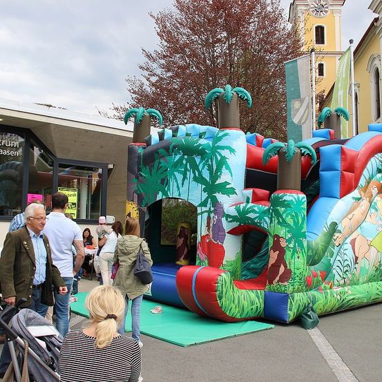 Ein bunter aufblasbarer Dschungel-Bouncer mit Rutsche ist draußen aufgestellt. Mehrere Leute sind um ihn herum, einschließlich eines Mannes, der einen Kinderwagen schiebt. Im Hintergrund befindet sich ein Gebäude mit Glasfenster und einem Glockenturm.