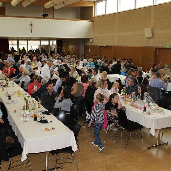 Eine große Gruppe von Menschen versammelt sich zum Essen in einem Saal. Sie sitzen um lange Tische, die mit weißer Tischdecke bedeckt sind. Einige Leute stehen in der Nähe der Tische, während andere sitzen. Der Saal hat Holzböden und Wände. Ein Kreuz ist an der Wand über dem Eingang angebracht.
