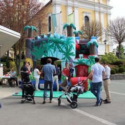 Eine Gruppe von Menschen mit Kinderwagen steht um ein riesiges aufblasbares Schloss mit Palmen im Freien.