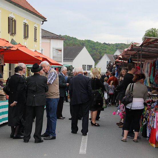 Eine Gruppe von Menschen versammelt sich auf einem Freiluftmarkt. Sie sind formell gekleidet, einige betrachten ausgestellte Gegenstände. Im Hintergrund befinden sich Häuser mit grünen Hügeln in der Ferne.