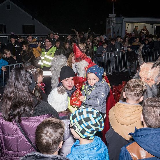 Eine Menschenmenge, einschließlich Kinder, versammelt sich um eine als Weihnachtsmann verkleidete Person. Einige tragen Winterkleidung und Mützen. Dahinter steht ein Metallzaun mit einem Fahrzeug.