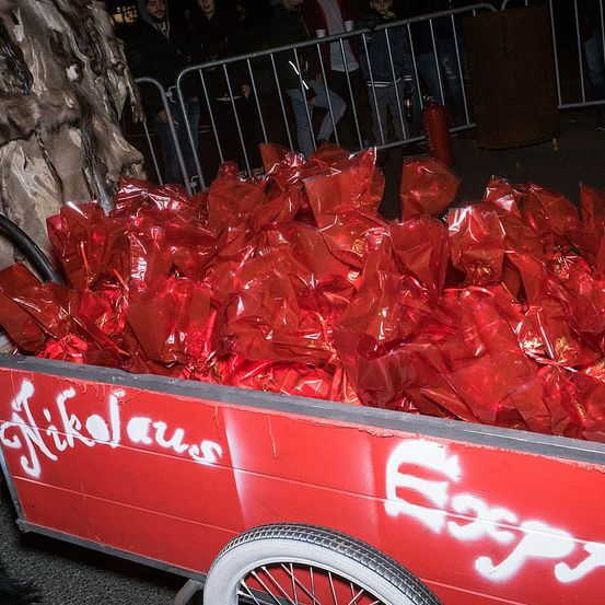 Ein roter Karren gefüllt mit roten Geschenktüten steht auf der Straße, Graffiti 'Nikolaus' und 'Epp' sind auf dem Karren. Mehrere Menschen stehen hinter einem Metallzaun.