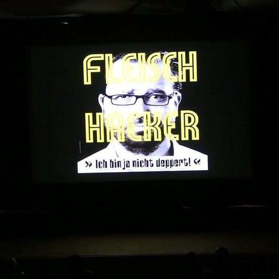Eine Leinwand zeigt ein Bild eines Mannes mit Brille, der ein weißes Hemd trägt, mit gelbem Text, der 'FLEISCH HACKER' über ihm und 'Ich bin ja nicht deppert!' darunter steht.