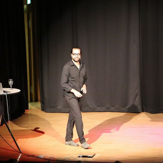 Ein Mann mit Brille und schwarzem Hemd steht auf der Bühne und hält eine Fernbedienung. Hinter ihm steht ein Tisch mit Laptop und Glas.