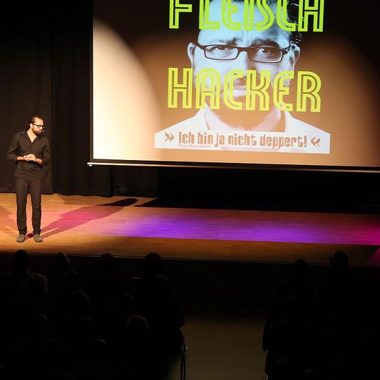 Ein Mann steht auf der Bühne und hält eine Präsentation, wobei hinter ihm eine große Leinwand einen Mann mit Brille zeigt und die Worte 'Fleisch Hacker'.