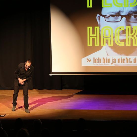 Ein Mann in schwarzer Kleidung steht auf der Bühne und schaut auf eine Leinwand, die ein Bild eines Mannes mit Brille zeigt. Das Bild sagt: 'Ich bin kein Hacker.'