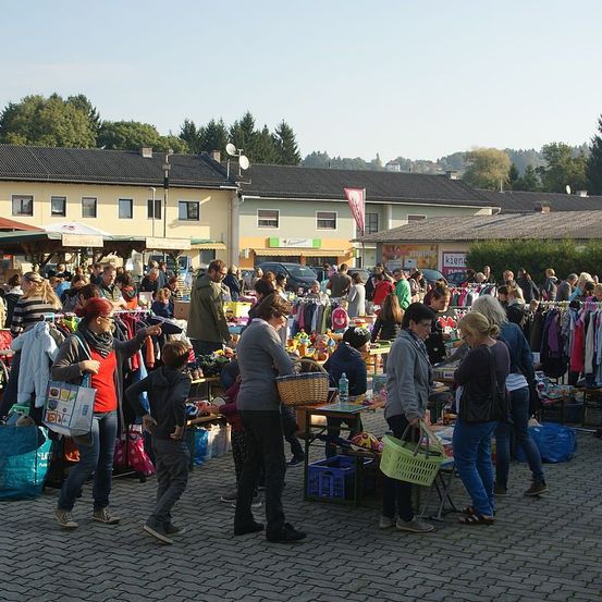 Ein geschäftiger Außenmarkt mit verschiedenen Ständen, die Kleidung, Spielzeug und mehr verkaufen. Menschen gehen herum, einige halten Einkaufstüten. Im Hintergrund Gebäude mit Geschäften und ein geparktes Auto.